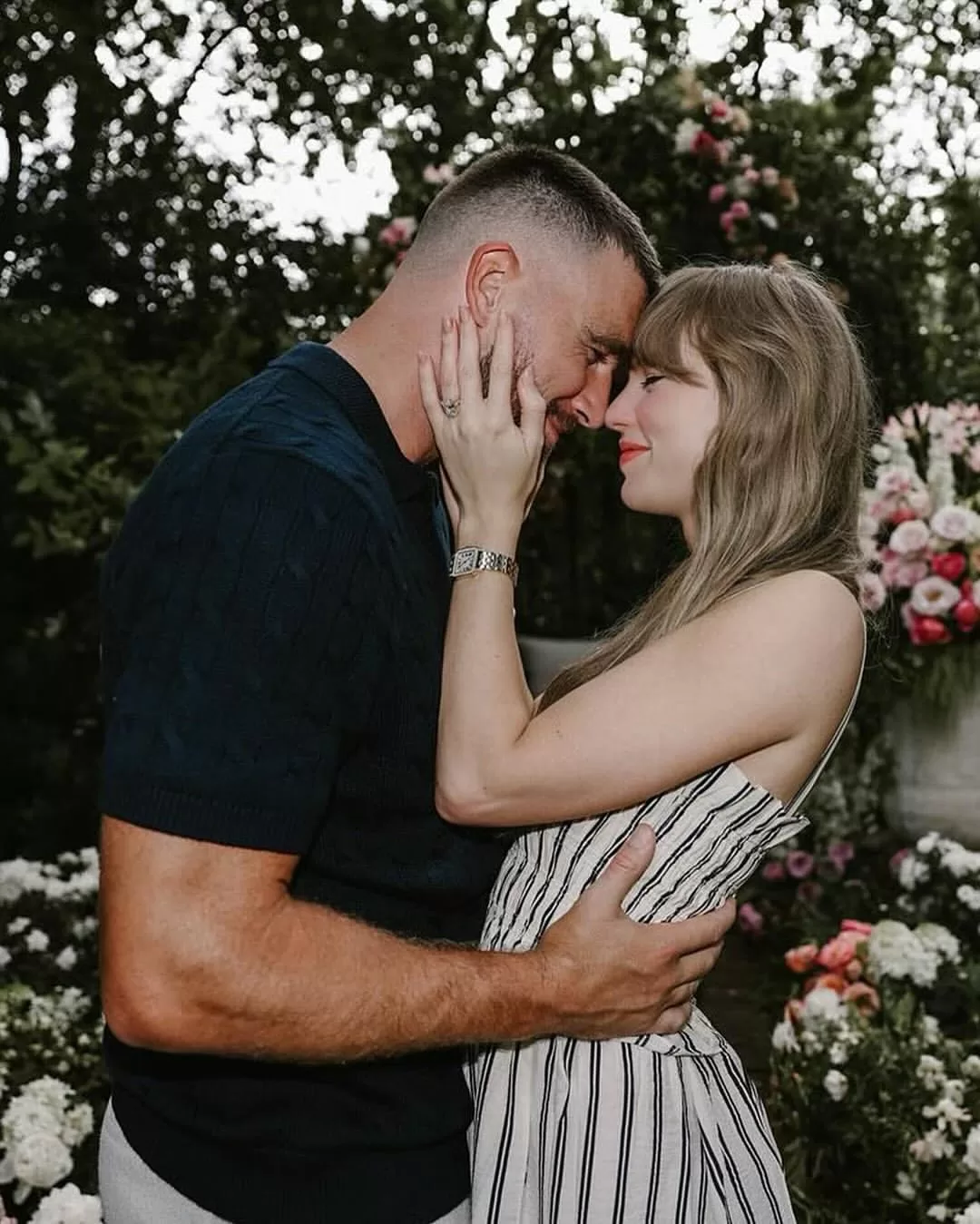 Taylor Swift and Travis Kelce smiling together under a floral arch during their engagement announcement, showcasing her Ralph Lauren dress and his classic style. | Aisle Society 