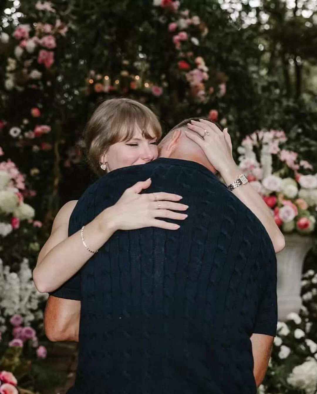Taylor Swift and Travis Kelce laughing and embracing during their engagement shoot, surrounded by romantic garden florals. | Aisle Society