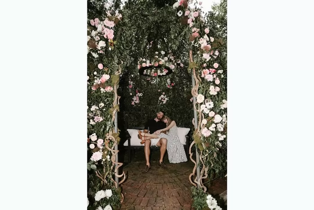 Taylor Swift and Travis Kelce smiling together under a floral arch during their engagement announcement, showcasing her Ralph Lauren dress and his classic style. | Aisle Society 